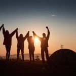 A silhouette of group people have fun at the top of the mountain near the tent during the sunset.