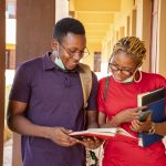 Two young African students wearing facemasks and holding books and phones at a campus